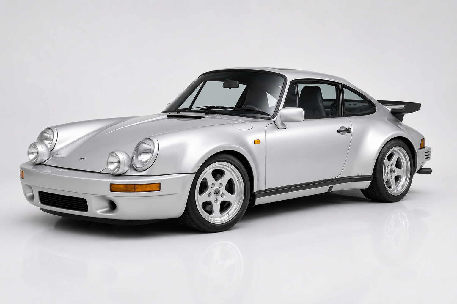 Silver RUF BTR in a studio setting, front three-quarter view showing the narrow-body 911/930 silhouette with RUF integrated fog and driving lights, RUF five-spoke wheels, side sills and a subtle rear spoiler — the foundational RUF turbo.