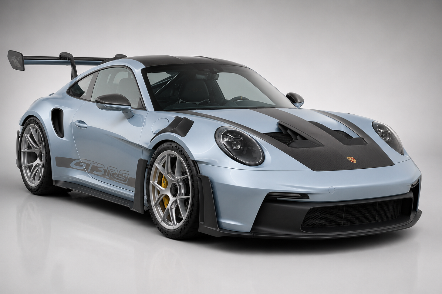 Light-blue Porsche 911 GT3 RS (992) in a grey studio, front three-quarter view showing the DRS-equipped swan-neck rear wing, exposed carbon bonnet with central duct and silver forged wheels.