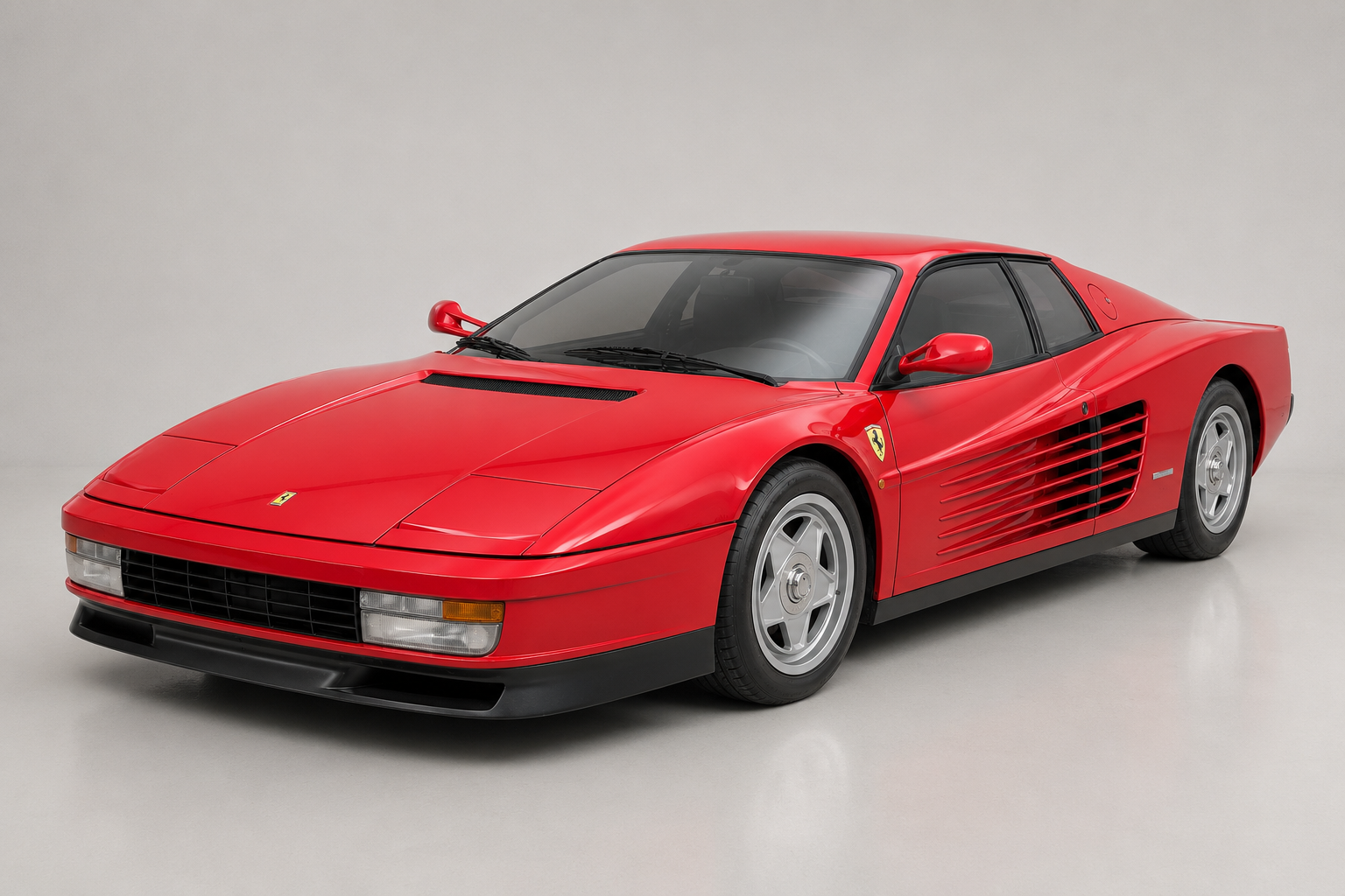 Red Ferrari Testarossa in a studio setting on a pale grey backdrop, front three-quarter view showing the retracted pop-up headlamps, bonnet vent, full-length side strakes feeding the side-mounted radiators, five-spoke alloy wheels and black lower sill trim.