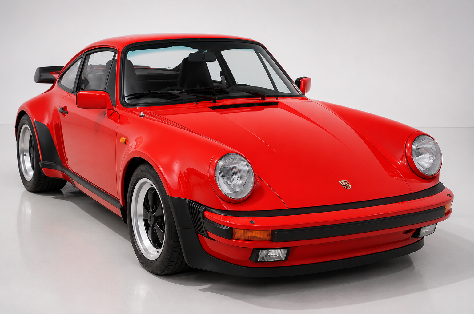 Red Porsche 911 Turbo (930) 3.3 coupe in a studio setting, front three-quarter view showing the flared rear arches, deep front spoiler with integrated fog lights, round headlamps and Fuchs-style alloy wheels.