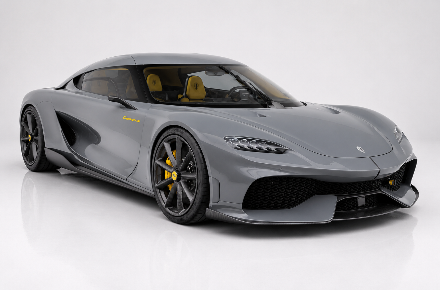 Grey Koenigsegg Gemera in a bright studio, front three-quarter view showing the four-door 'Mega-GT' silhouette with yellow 'Gemera' script on the front fender, wraparound cabin glass with yellow leather interior visible, deep front splitter, side intakes and multi-spoke black wheels with yellow brake calipers.