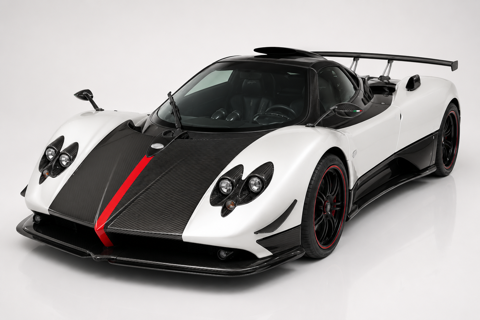 White and exposed-carbon Pagani Zonda Cinque in a studio setting with red centre-stripe accents, front three-quarter view showing the carbon front section, quad-round LED headlight clusters, low splitter and multi-spoke wheels with red pinstriped rims.