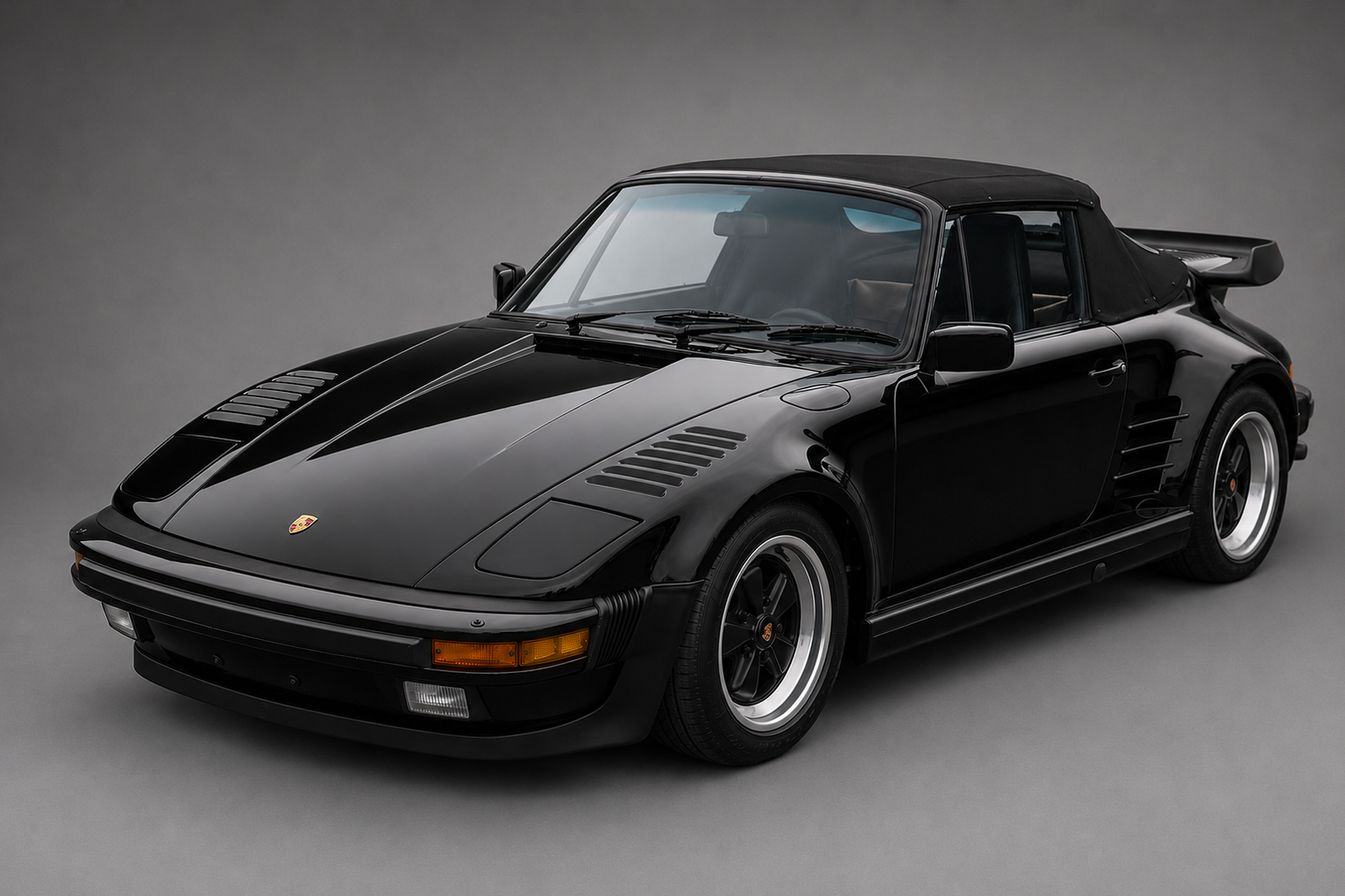 Studio three-quarter front view of a black Porsche 911 Turbo Flachbau Cabriolet (930) with roof raised, flat-nose front end with pop-up headlights closed, side louvres and black five-spoke wheels, on a plain grey backdrop.
