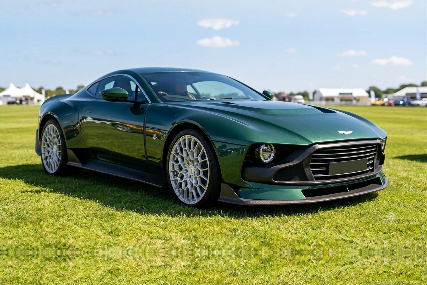 Dark green Aston Martin Valour coupe on grass at an outdoor event, front three-quarter view showing the retro-inspired coach-built bodywork, wide mesh grille, round headlamps and gold-toned multi-spoke alloy wheels.