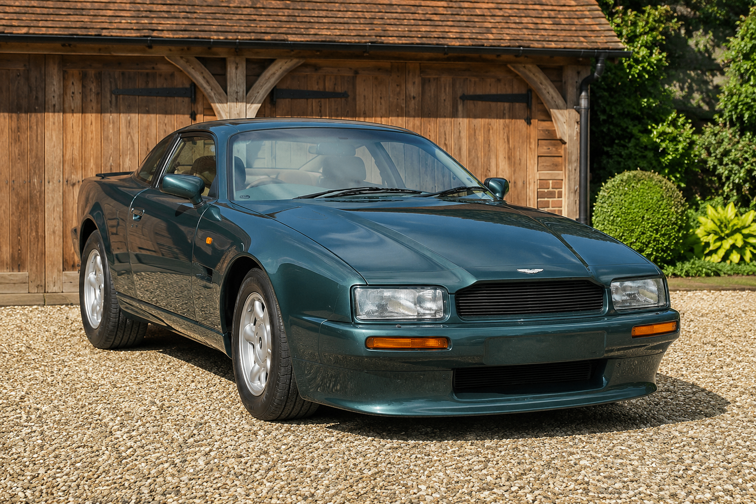 Aston Martin Virage Coupé in dark metallic green, front three-quarter view on gravel in front of a wooden garage, showing the body-colour bumpers, cream leather interior and multi-spoke alloy wheels.