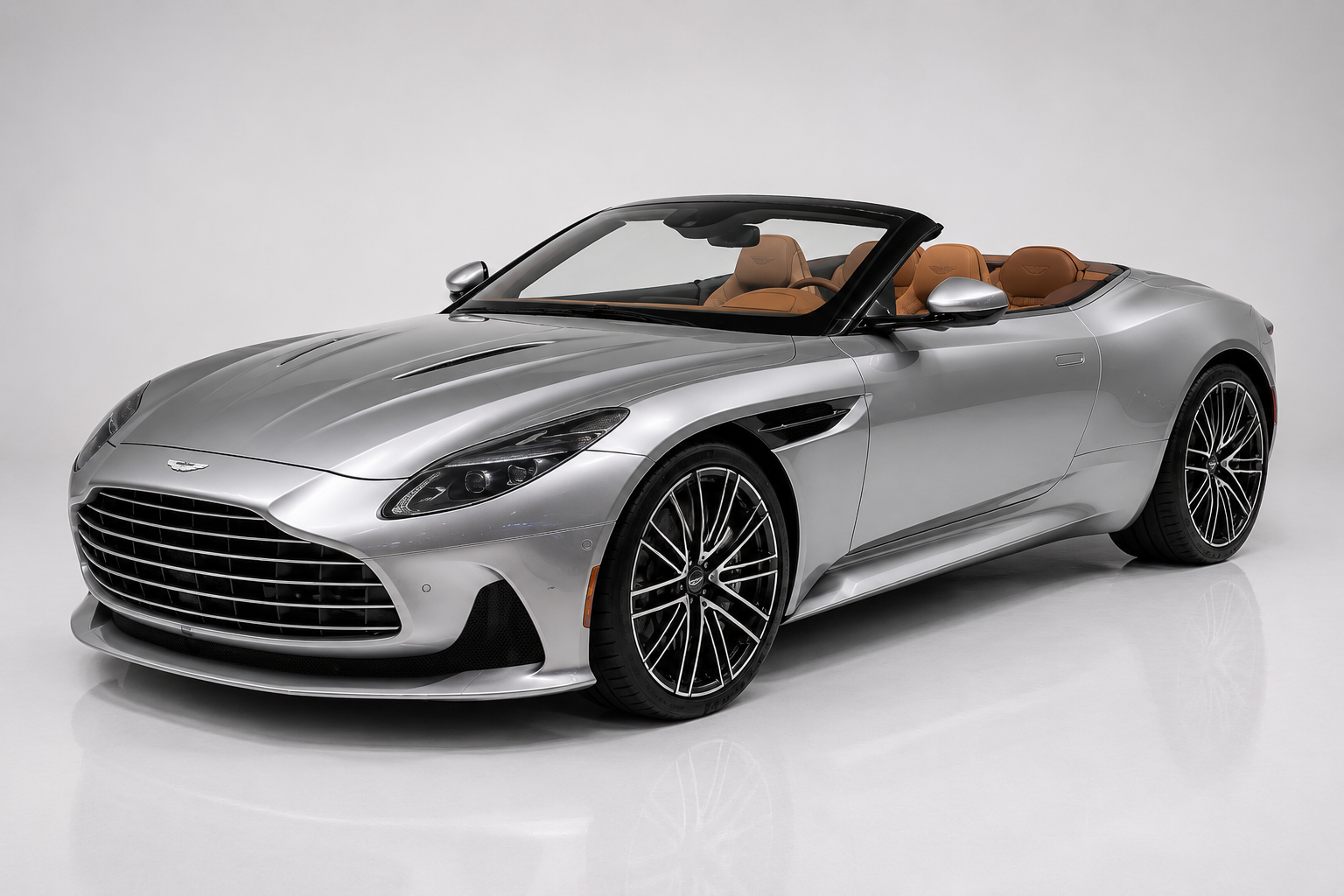 Silver Aston Martin DB12 Volante convertible with roof lowered, in a bright studio, front three-quarter view showing the enlarged grille, sharp full-LED headlamps, sculpted bonnet, wide front air intakes and multi-spoke black-and-machined alloy wheels, with tan leather interior — the first Aston with the brand's new winged logo.