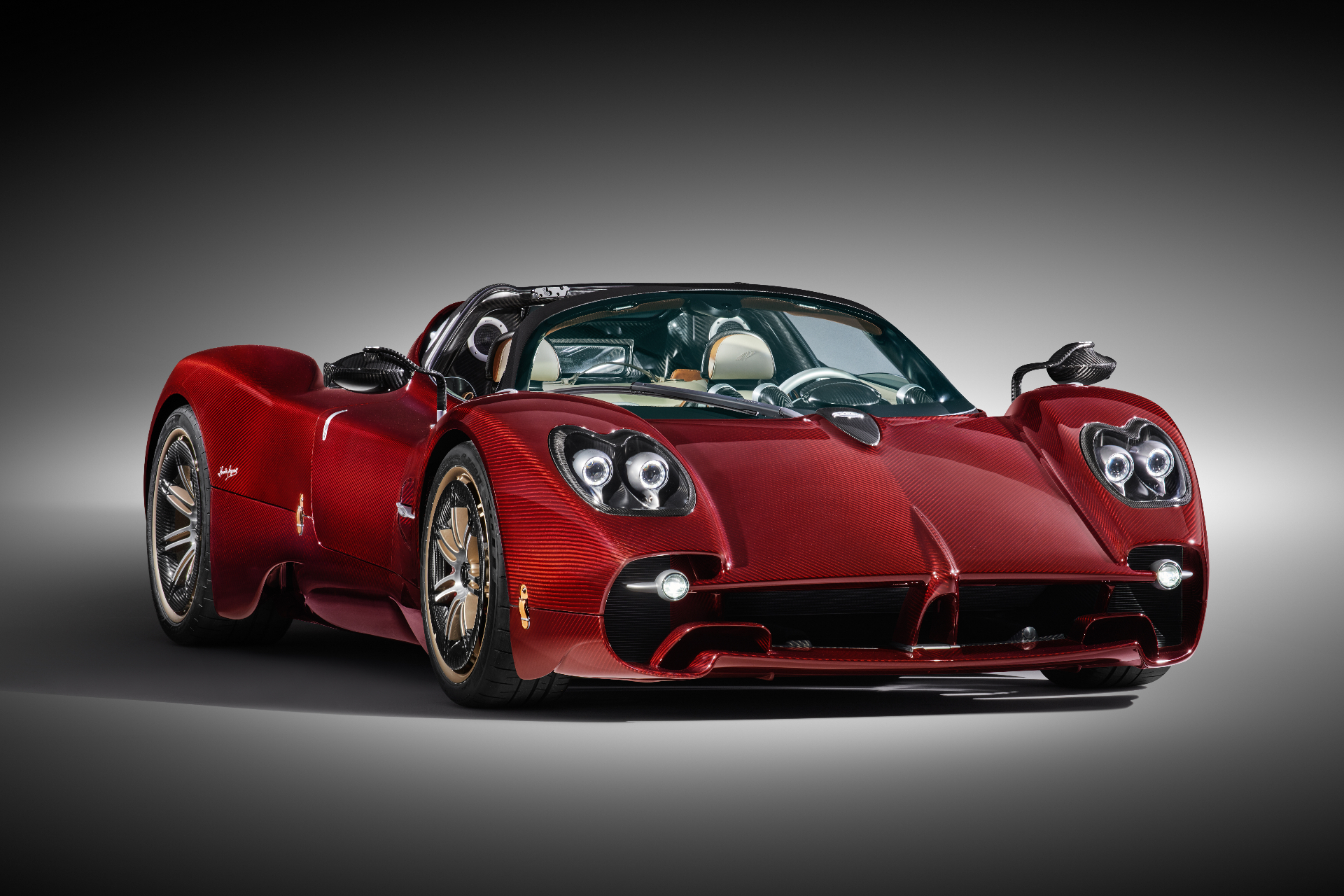 Red carbon-fibre Pagani Utopia in a studio, front three-quarter view showing the quad-headlamp clusters, exposed carbon weave, gold-accented wheels, open-top cabin with cream leather seats and the sculpted rear buttresses.
