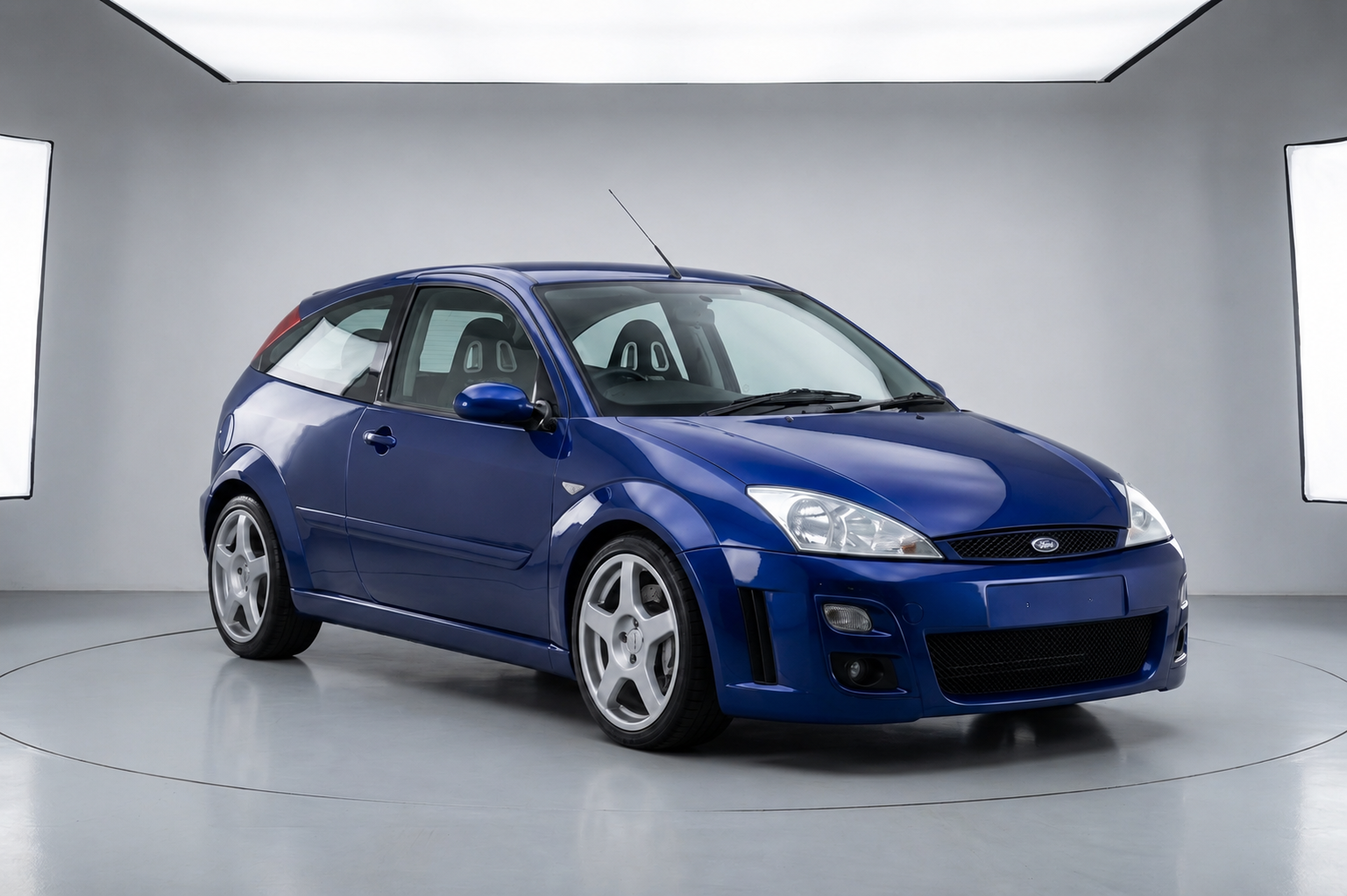 Ford Focus RS Mk1 in Imperial Blue on a studio turntable, showing the three-door bodyshell, blistered front and rear arches, deep front bumper with mesh grille and centre-lock-style five-spoke OZ Racing alloy wheels — the 2002–2003 first-generation Focus RS in canonical launch specification.
