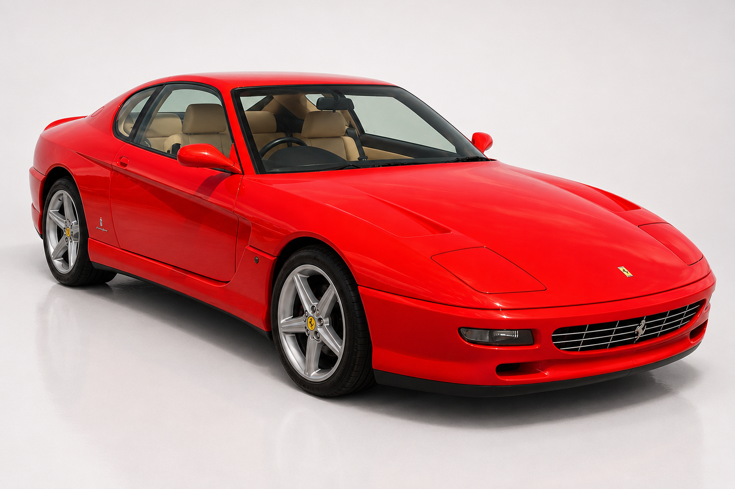 Red Ferrari 456 GT coupe in a bright grey studio setting, front three-quarter view showing the smooth Pininfarina bodywork, retractable pop-up headlamps, oval lower grille, tan leather interior and silver five-spoke alloy wheels.