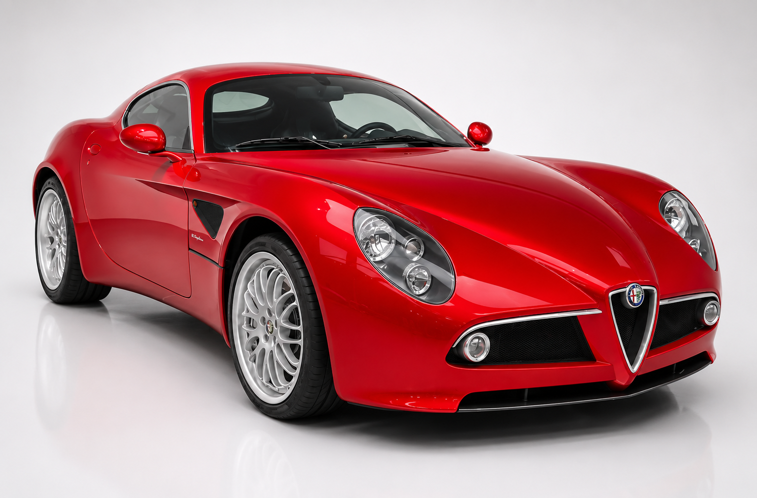 Red Alfa Romeo 8C Competizione coupé in a bright studio, front three-quarter view showing the shield grille, twin round headlights under clear covers, sculpted bonnet, side gill vent behind the front wheel and silver multi-spoke alloy wheels.