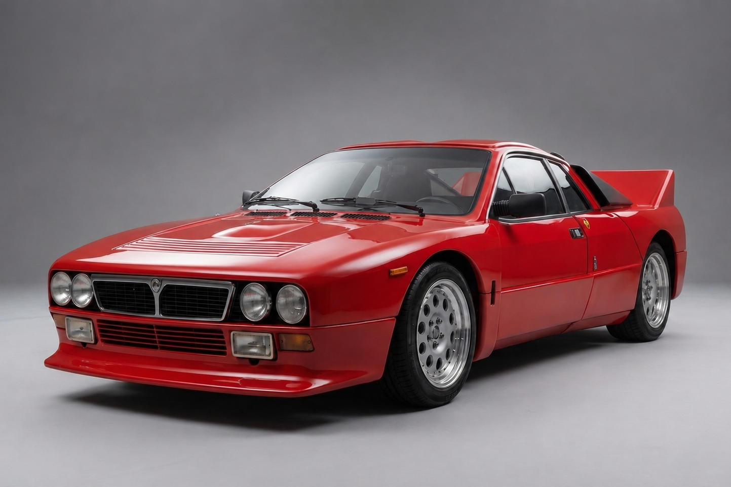 Lancia Rally 037 Stradale in Rosso Corsa, front three-quarter studio view showing the Pininfarina wedge nose, exposed quad round headlamps, deep front air dam, silver multi-hole alloy wheels and the tall rear wing rising above the flying-buttress rear haunches — the road-going homologation for Lancia's Group B rally weapon.