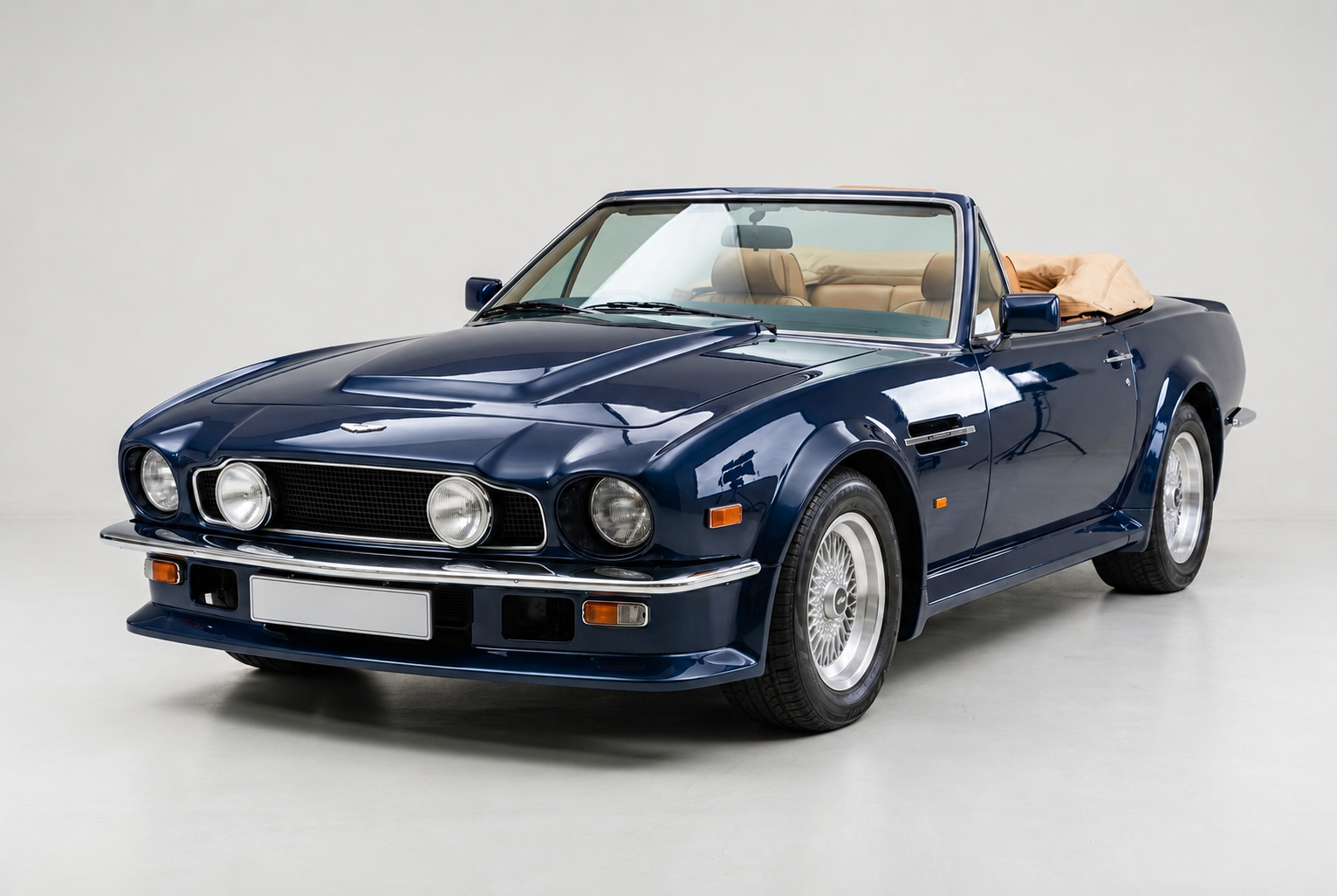 Dark blue Aston Martin V8 Vantage Volante convertible with tan leather interior and folded soft-top, front three-quarter studio view on a plain pale grey backdrop showing the blanked-off grille, quad round headlamps, chrome bumpers with side-lamp pods and multi-spoke alloy wheels.
