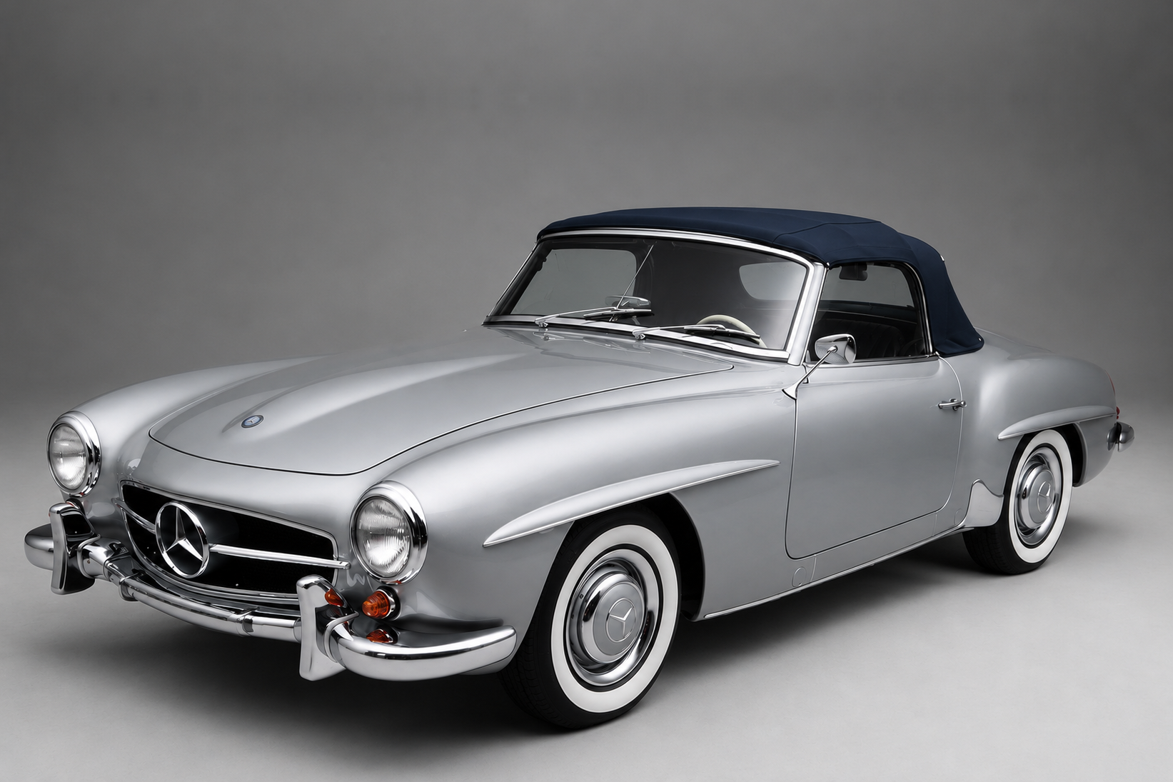 Silver Mercedes-Benz 190 SL roadster with dark blue soft top raised in a plain grey studio setting, front three-quarter view showing the chrome grille with central three-pointed star, chrome bumper with over-riders, whitewall tyres and body-colour steel wheels with chrome hub caps.